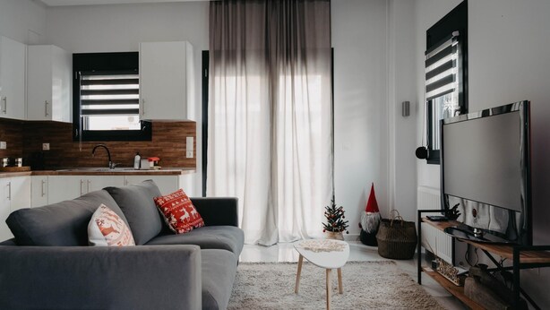 Una sala moderna con un sofá gris, una pequeña mesa de centro blanca, un televisor sobre un mueble de madera, y una zona de cocina con armarios blancos y un protector de salpicadero de madera.