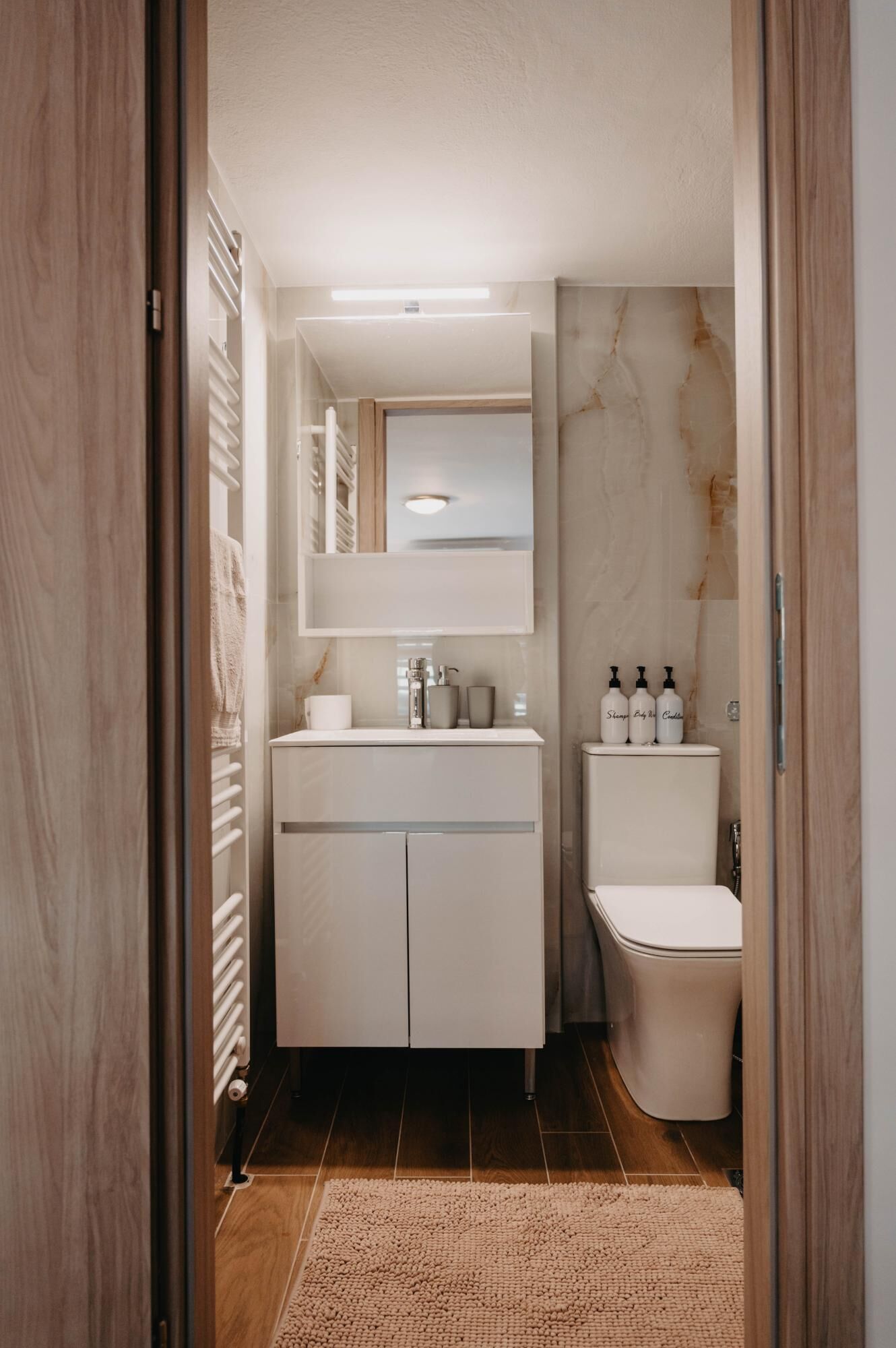 Un baño moderno con tocador blanco, espejo y retrete. Hay artículos de aseo en el lavabo y un perchero para toallas.
