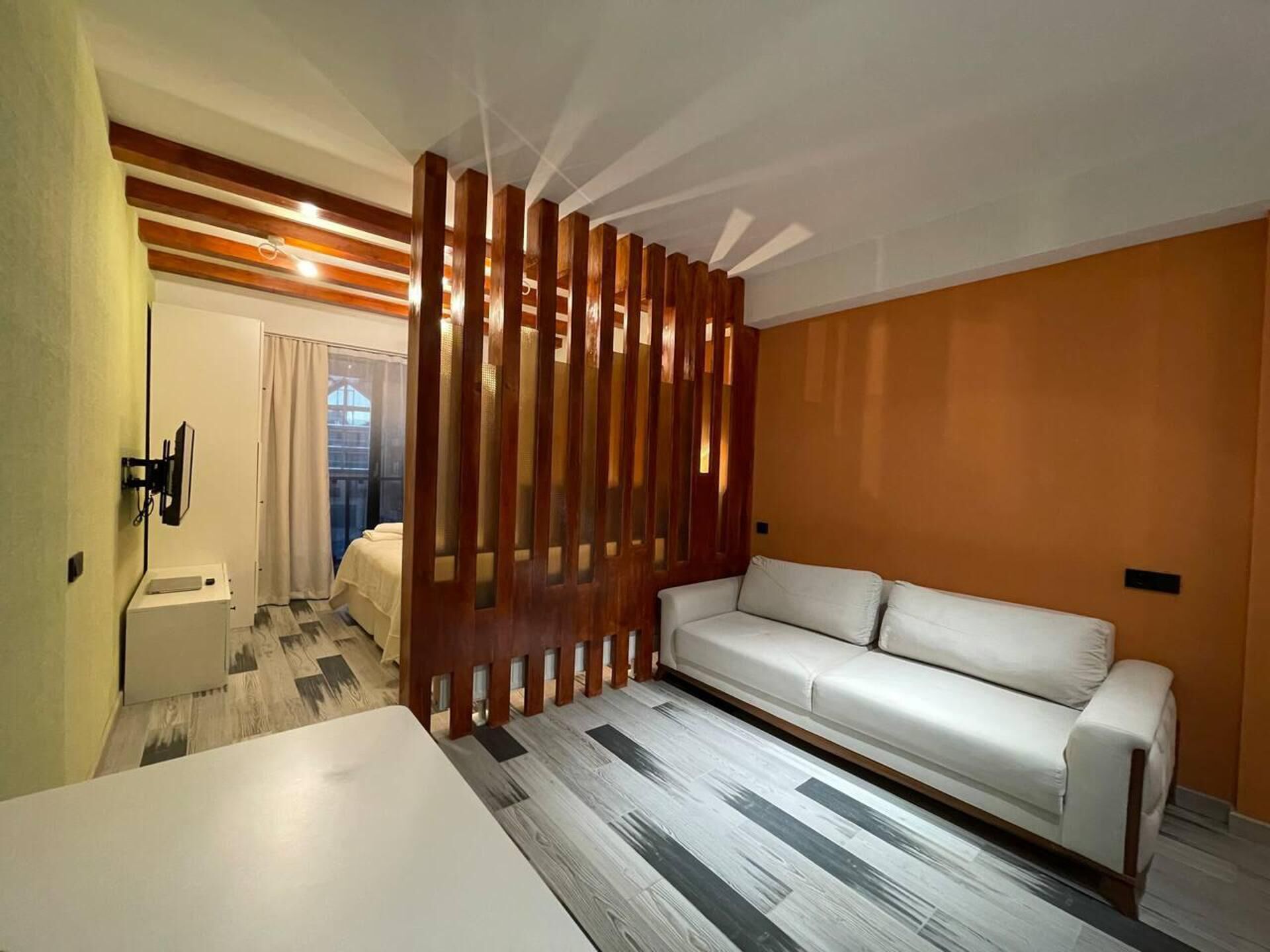 A modern hotel room with a white sofa, a wooden partition, a small bedside table, and a view of the outdoors.