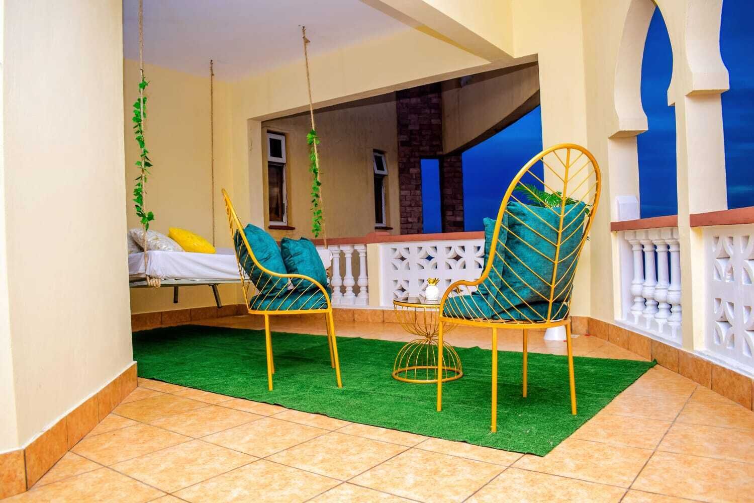 Una habitación con dos sillas metálicas amarillas y cojines verdes, una mesita con una silla amarilla, y una cama con ropa de cama blanca y amarilla.