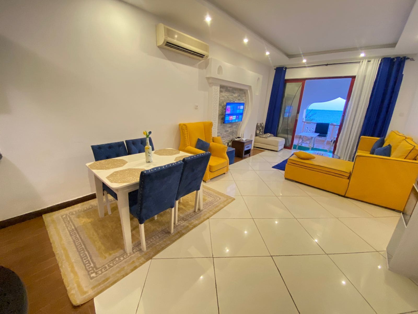 A modern living room with a dining area, yellow sofas, a flat-screen TV, and a view of the sea.