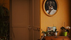 A room with a wooden dresser, a table with a checkered tablecloth, a chair, a door, a framed picture on the wall, and potted plants.