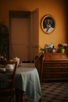 Um quarto com cômoda de madeira, uma mesa com toalha quadriculada, uma cadeira, uma porta, um quadro na parede e plantas em vasos.