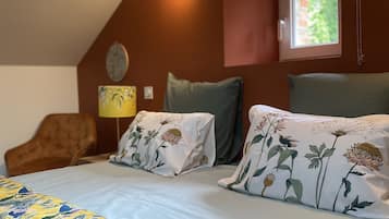 Quarto com teto inclinado, cama com roupa de cama florida, duas almofadas com estampas florais, mesa de cabeceira com abajur e janela com vista para área verde.