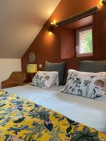 Un dormitorio con techo inclinado, una cama con ropa de cama floral, dos almohadas con diseños florales, una mesita de noche con una lámpara y una ventana con vistas a follaje.