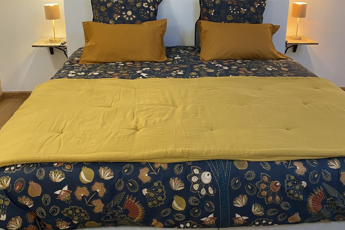 Un lit bien fait, avec une couette à motifs floraux et deux coussins de couleur moutarde, encadré de deux lampes de chevet posées sur des tables de nuit assorties.