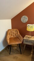 Quarto de canto com poltrona estofada, abajur com cúpula florida e um prato decorativo na parede.