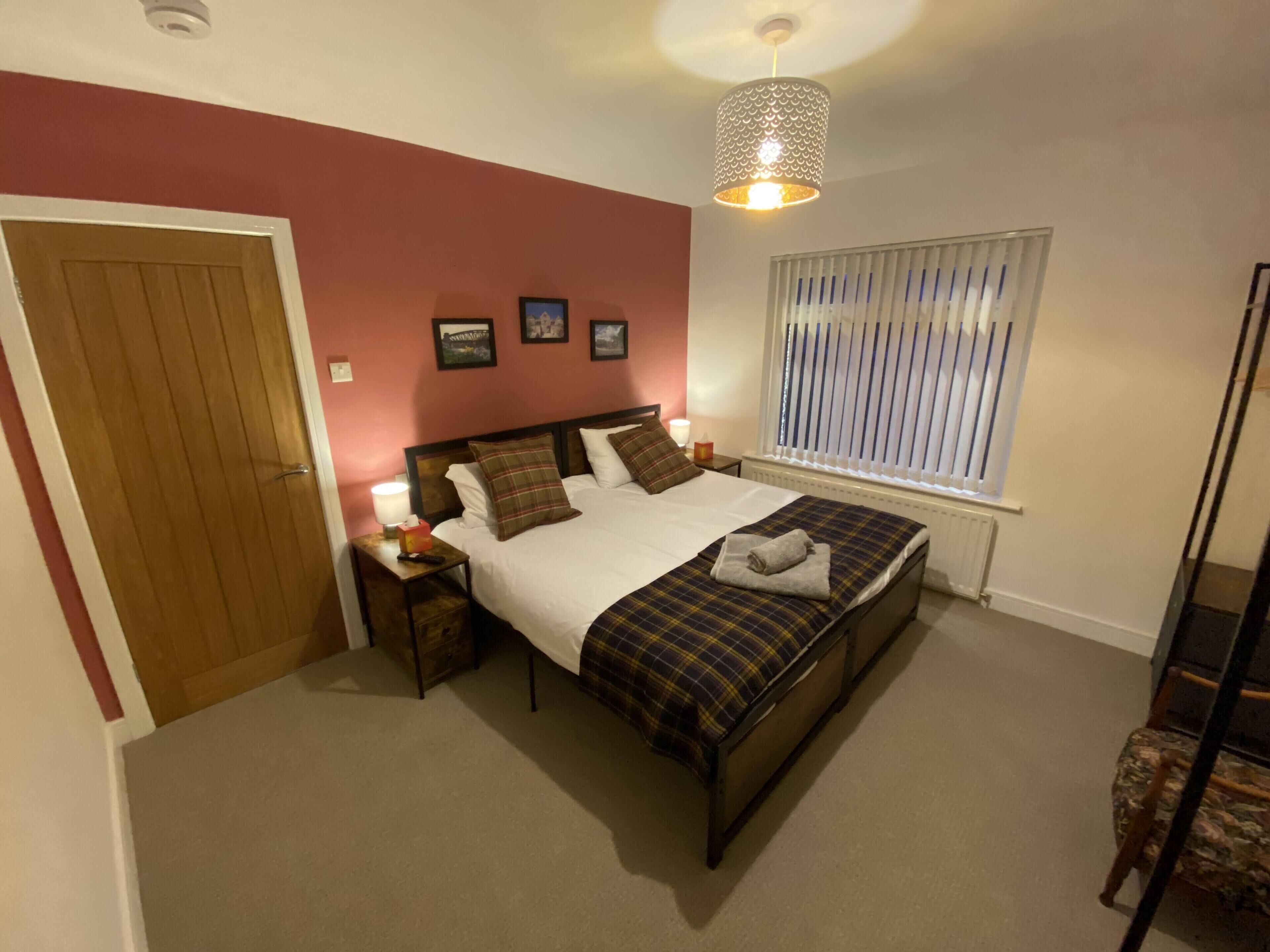 A bedroom with a bed, a nightstand, a lamp, a window with blinds, and a door.