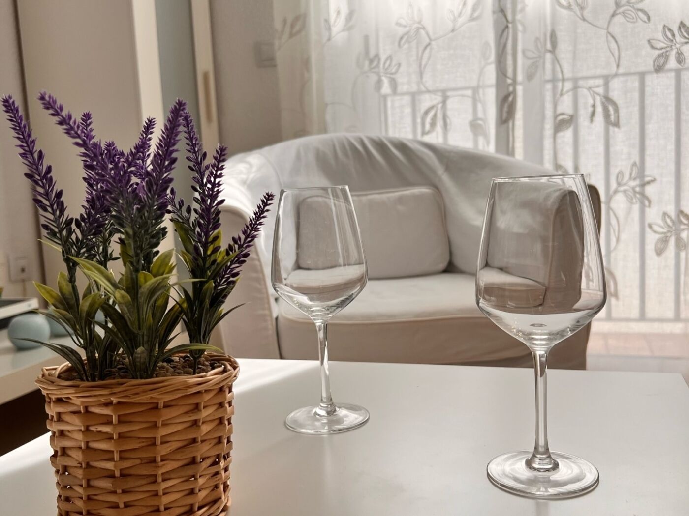 Een kamer met een witte tafel, twee lege wijnglazen en een geweven mand met paarse bloemen.