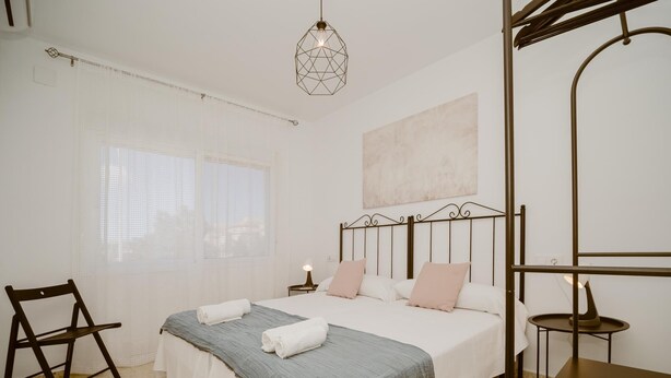 Un dormitorio con cama, una silla, una mesa, una lámpara colgante y una ventana con cortinas traslúcidas.