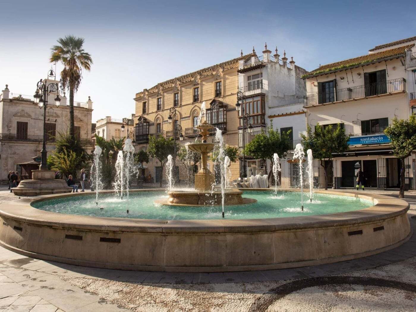 A public square with a central fountain, surrounded by historic buildings and a palm tree.