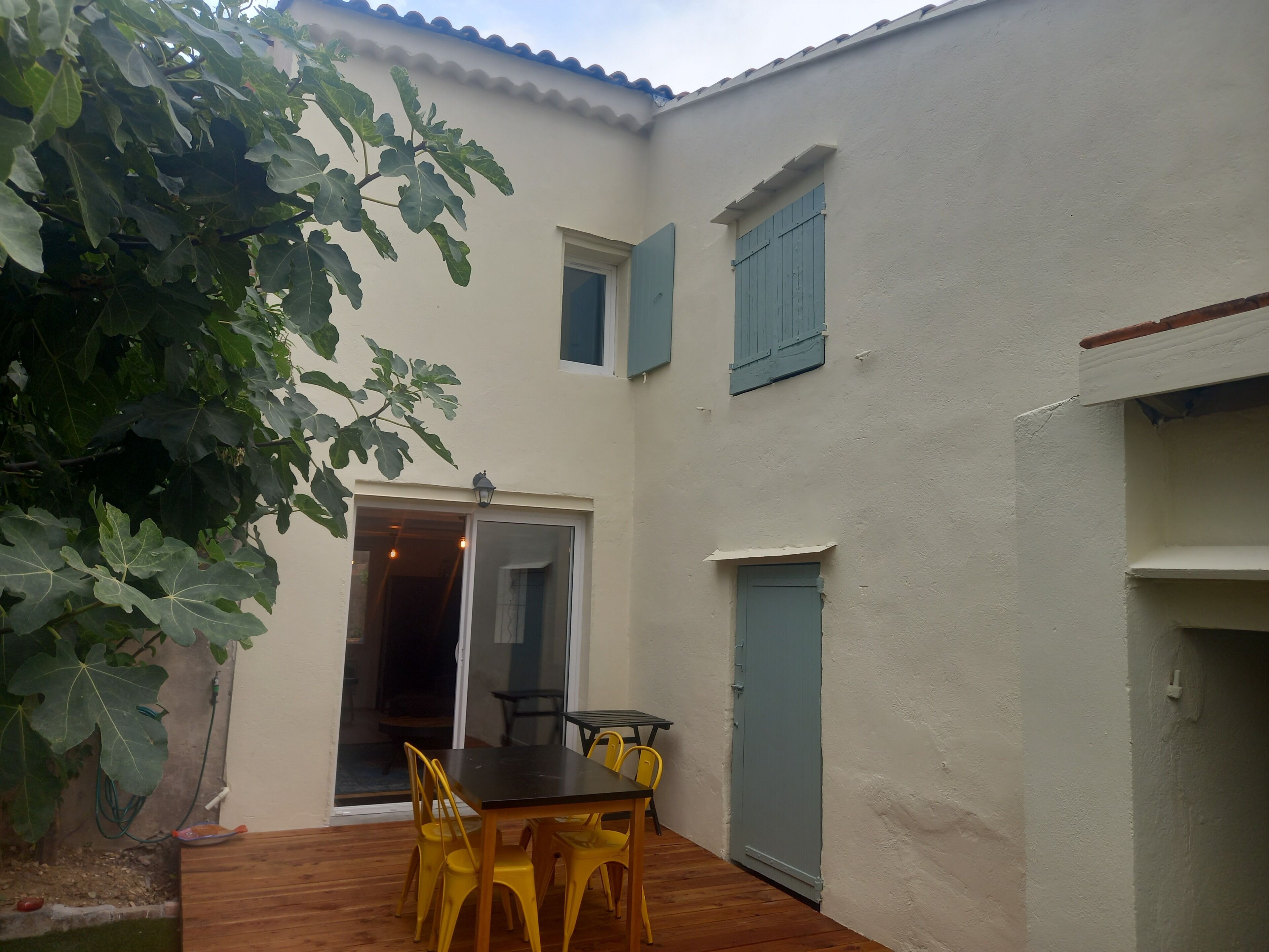 Un patio con una mesa de madera y sillas amarillas, una puerta corrediza de vidrio y un ventanal con contraventanas azules.