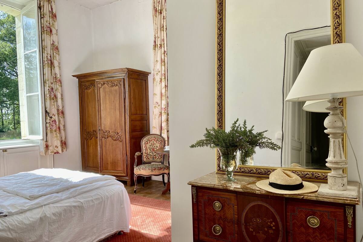 A bedroom with a bed, a wooden wardrobe, a chair, a vanity table with a lamp, and a mirror.