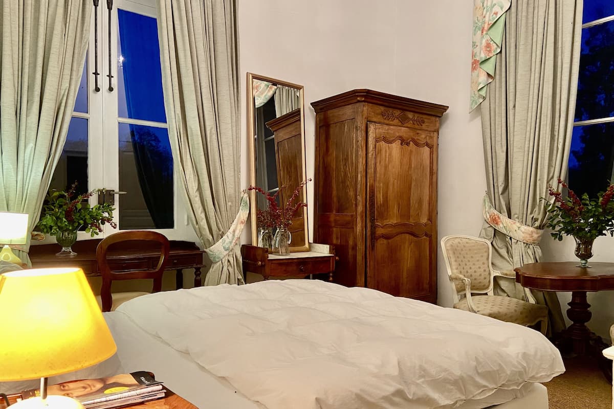 A hotel room with a bed, a wooden wardrobe, a desk with a lamp, and a chair.