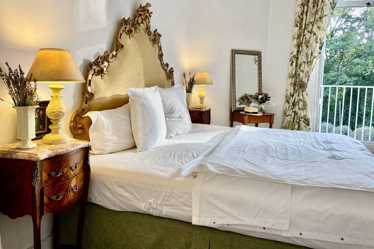 A neatly made bed with white linens, two bedside tables with lamps, a mirror, and a view of greenery outside.