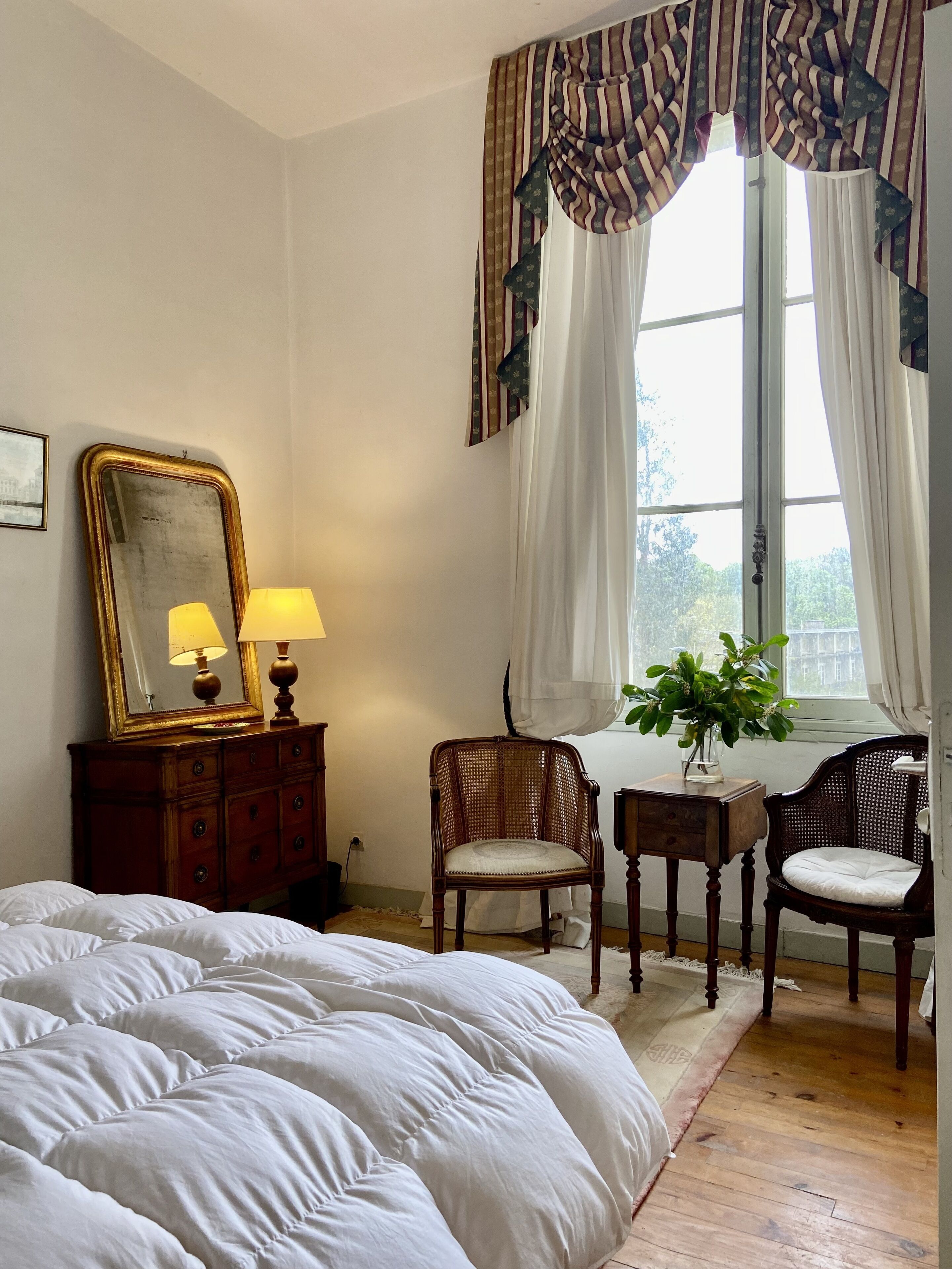 Un dormitorio con una cama grande, un aparador con espejo, dos sillas y un ventanal con cortinas.