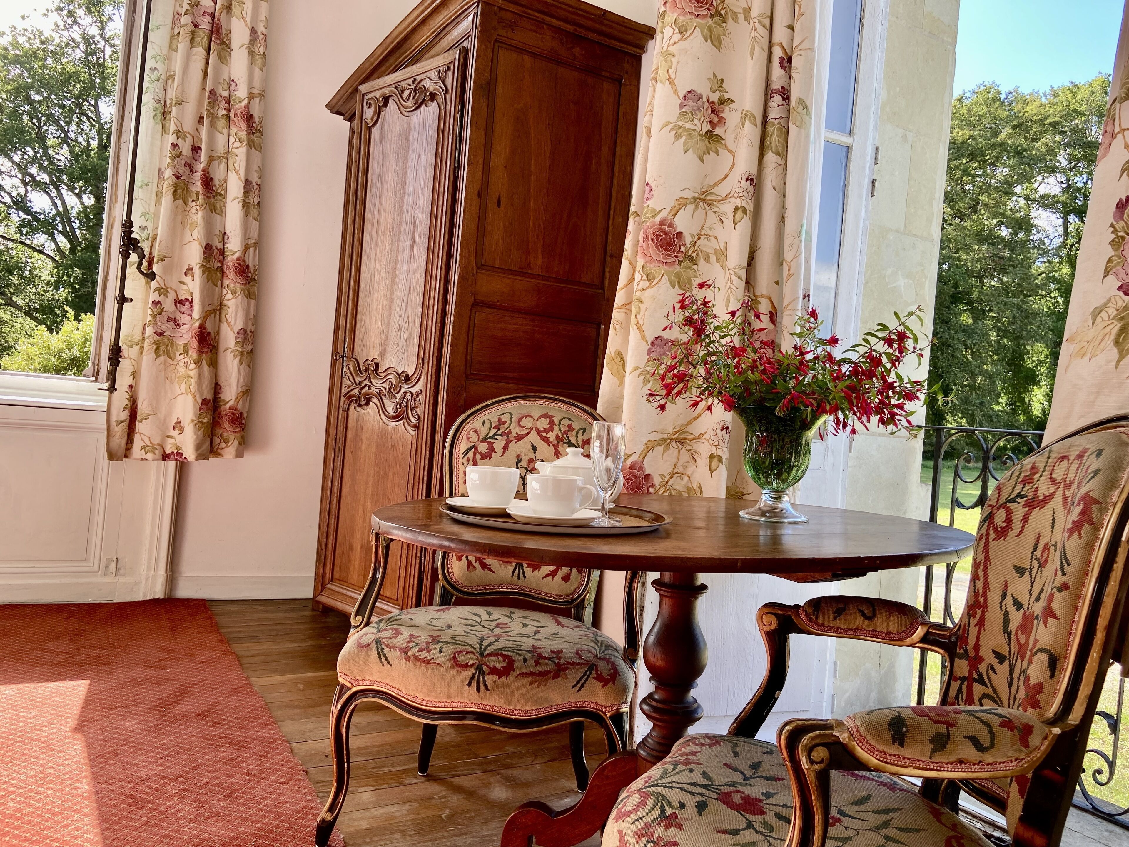 A room with a wooden cabinet, two chairs with floral upholstery, a round table with a vase of flowers, and a window with floral curtains.