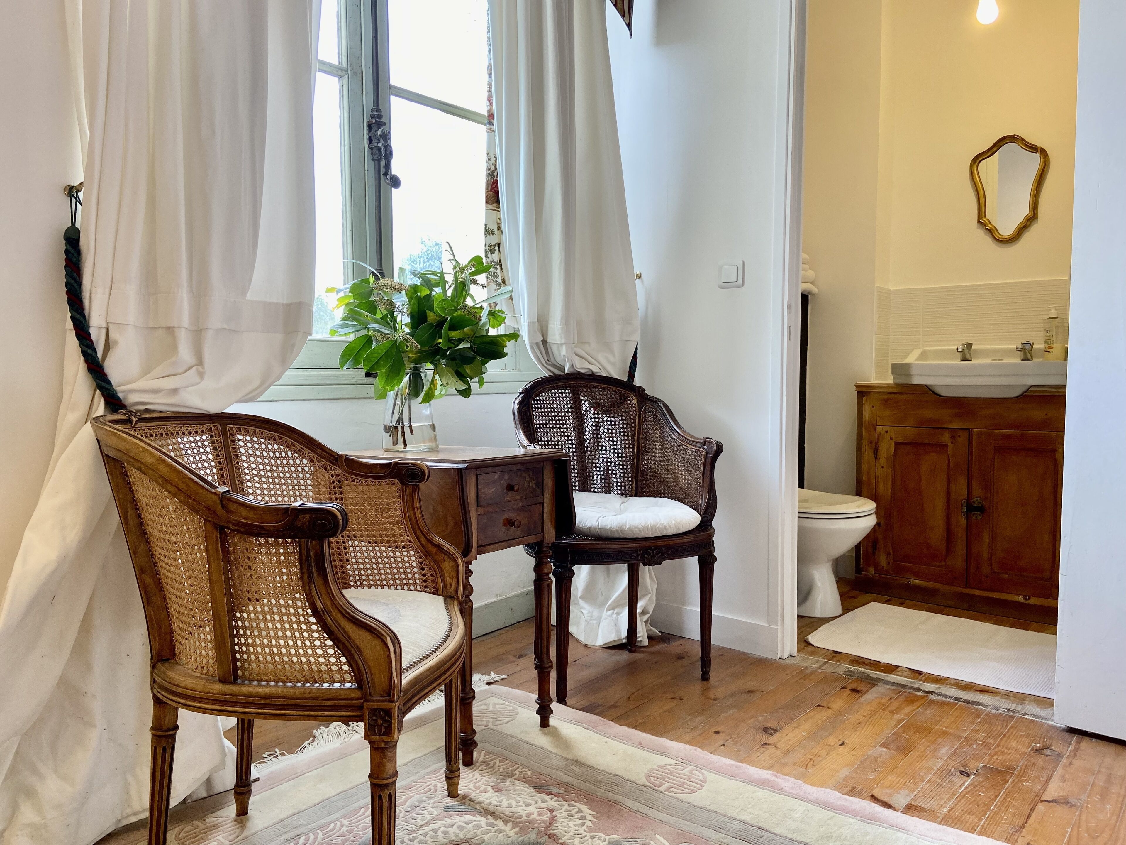 A bathroom with wooden cabinets, a sink, and a toilet. There are two wicker chairs and a small table with a vase of flowers.