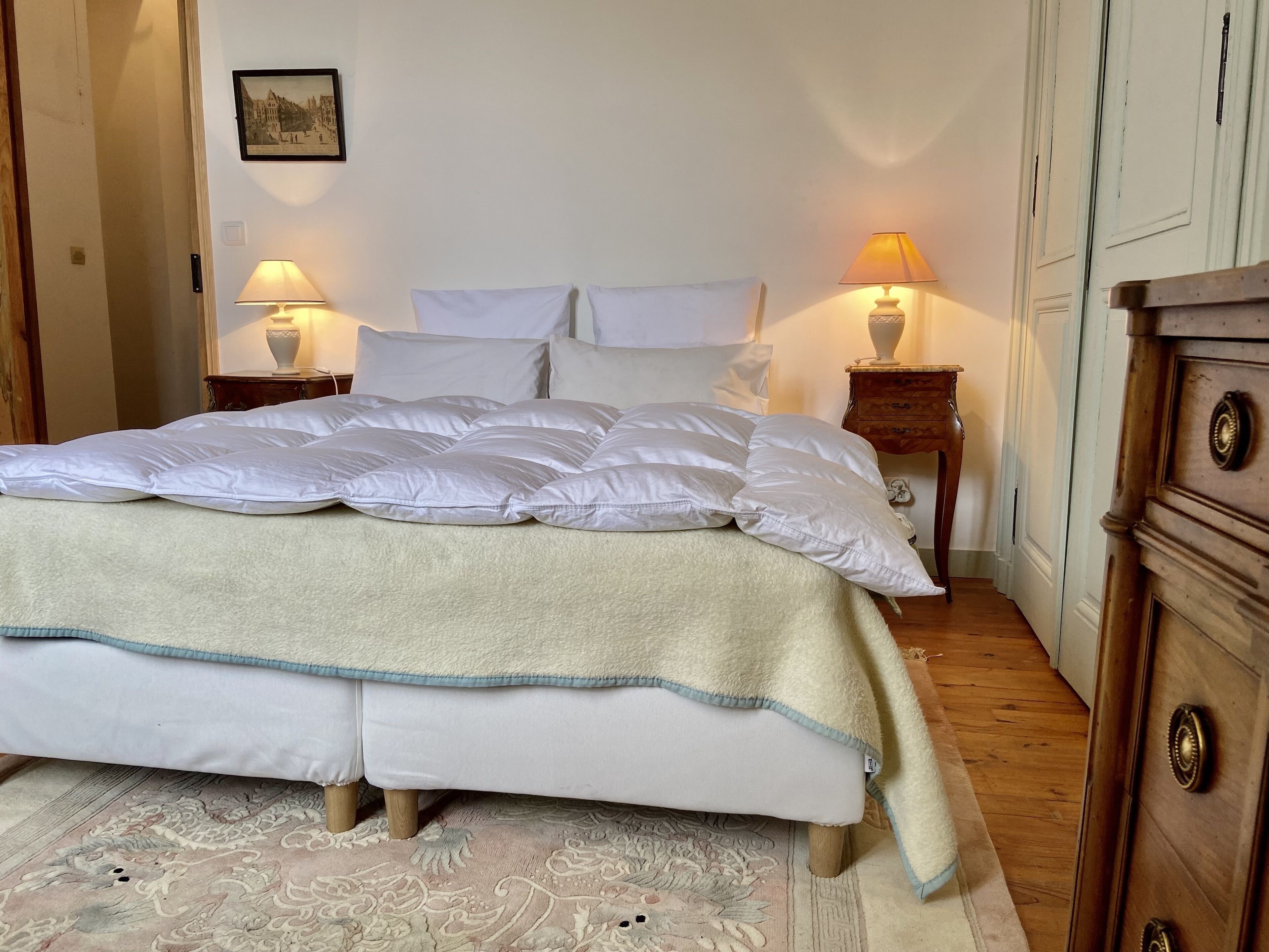 A neatly made bed with a white duvet and pillows, two bedside lamps, and a wooden dresser to the right.