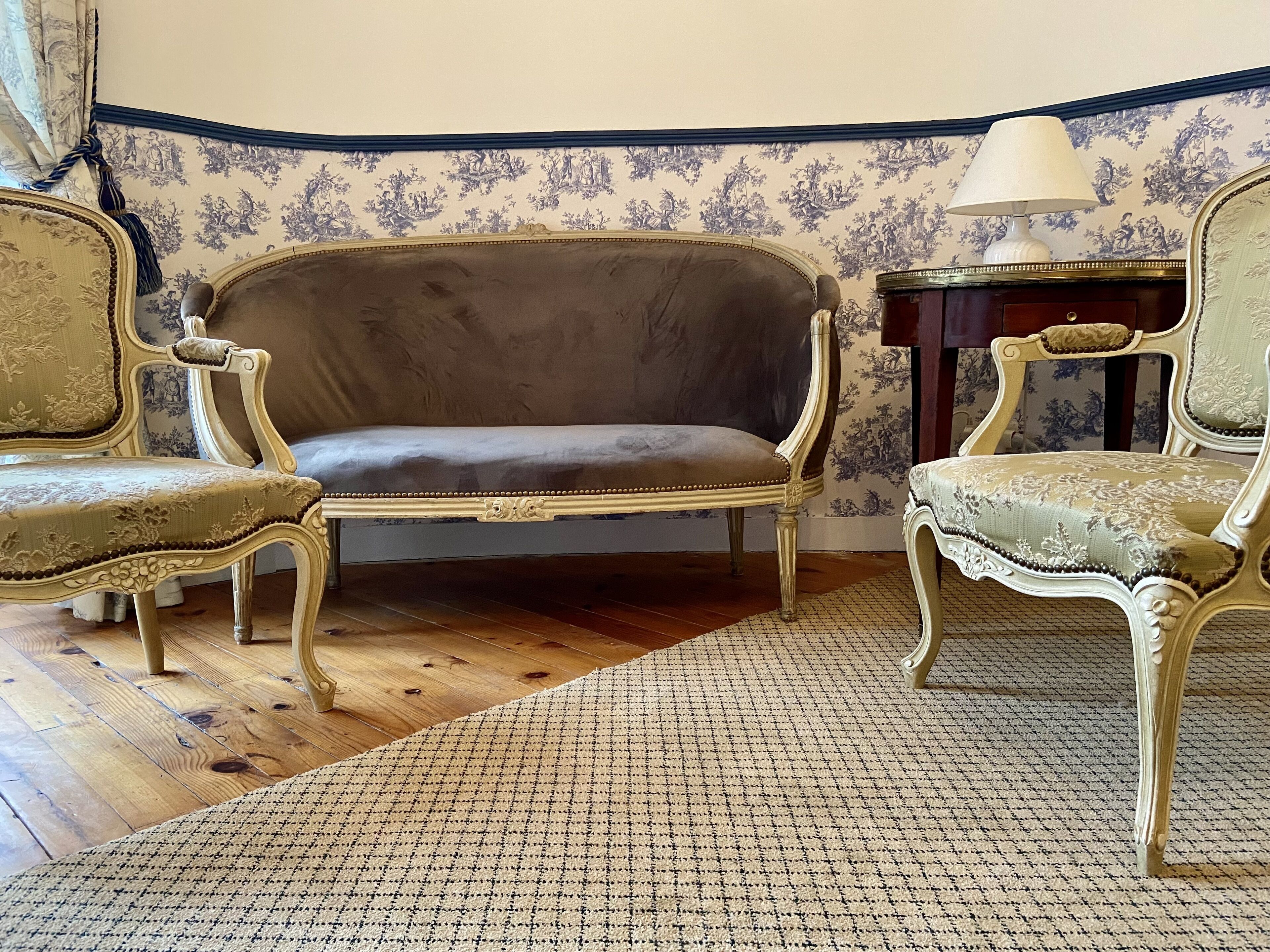 A room with a patterned wallpaper, a wooden floor, and two ornate chairs with a table and a lamp.