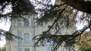 Un edificio grande e histórico con fachada simétrica, ventanas amplias y una entrada central. En primer plano, hay dos sillones de madera con cojines verdes.