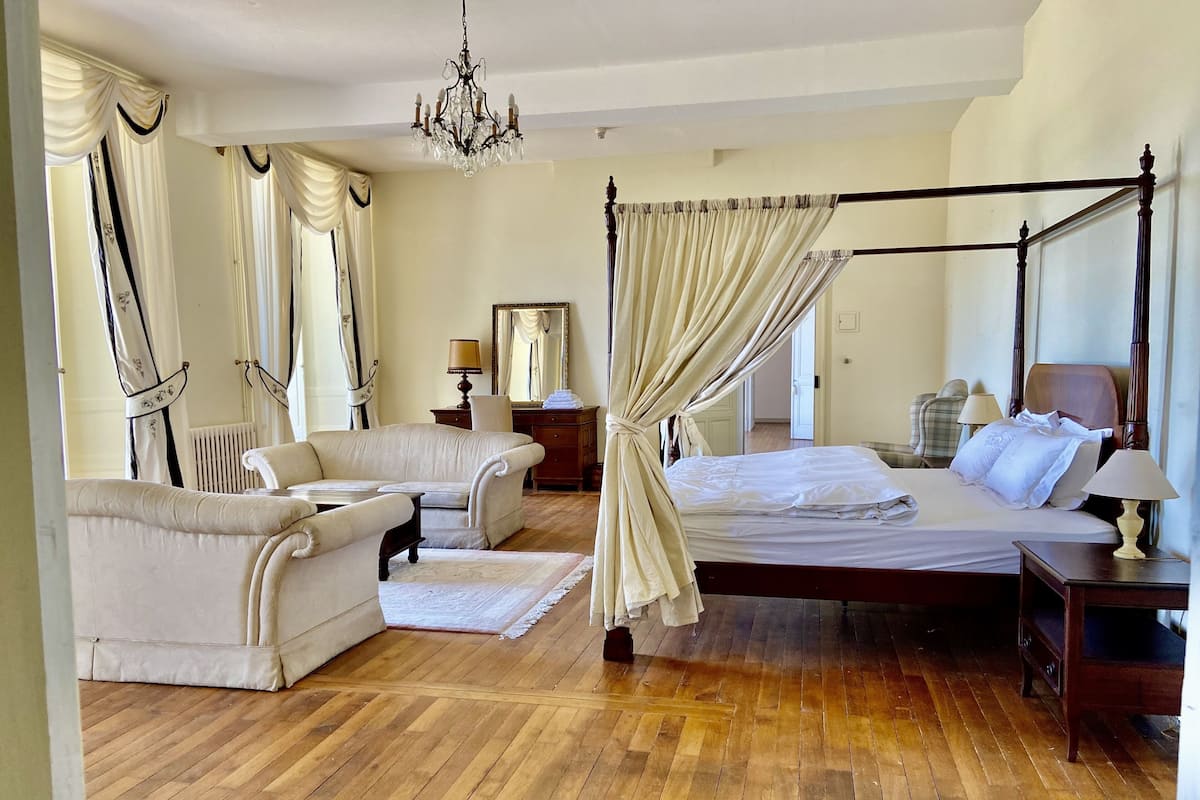 A spacious bedroom with a four-poster bed, a sitting area with a sofa and armchairs, a wooden floor, and a chandelier.