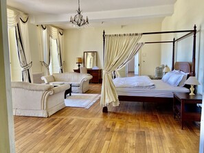 Ein geräumiges Schlafzimmer mit Himmelbett, einer Sitzecke mit Sofa und Sesseln, Holzboden und einer Kandelaberleuchte.