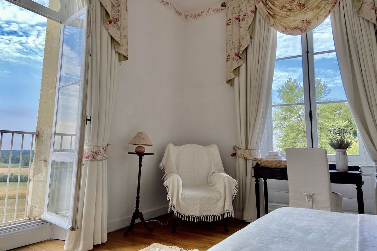 A room with a white armchair, a small table with a lamp, and a view of the outdoors through large windows.