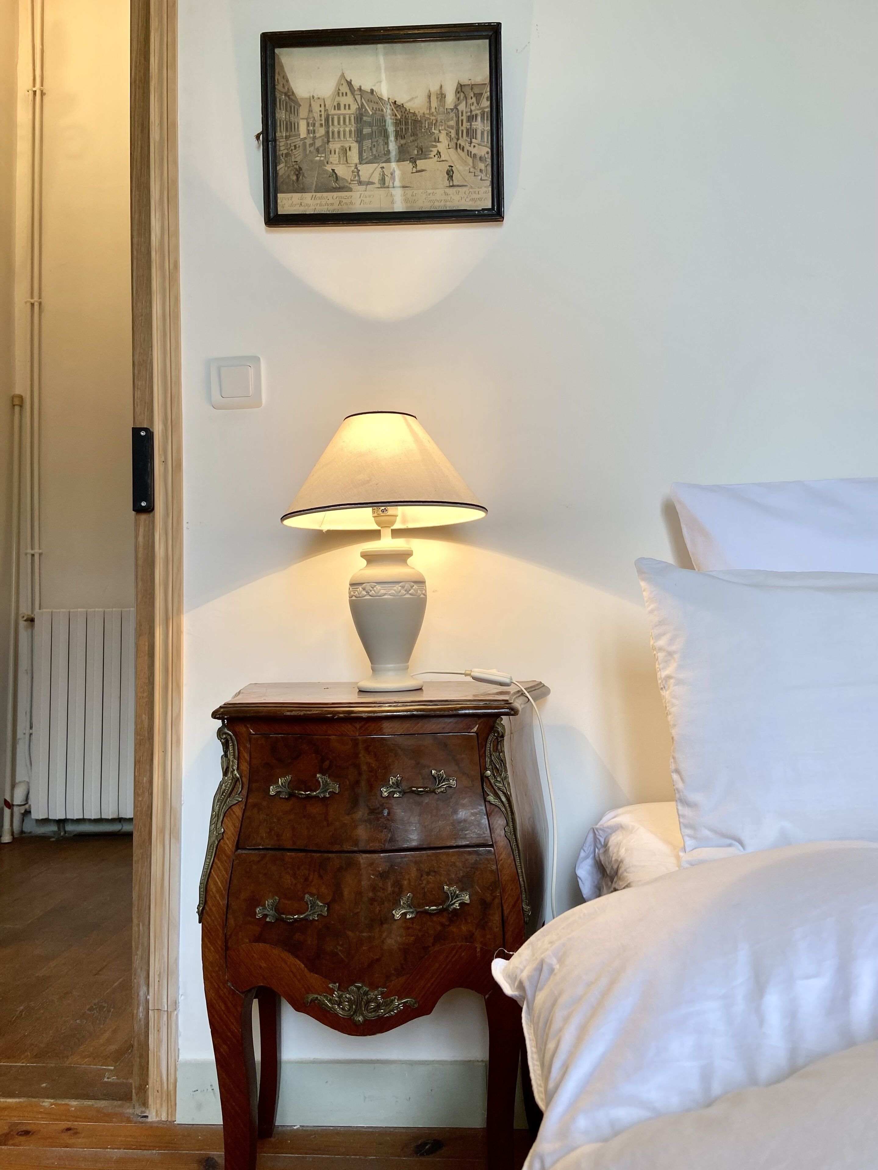Een netjes ingerichte slaapkamer met een bed, nachtkastje met lamp, een dressoir en een ingelijst kunstwerk aan de muur.