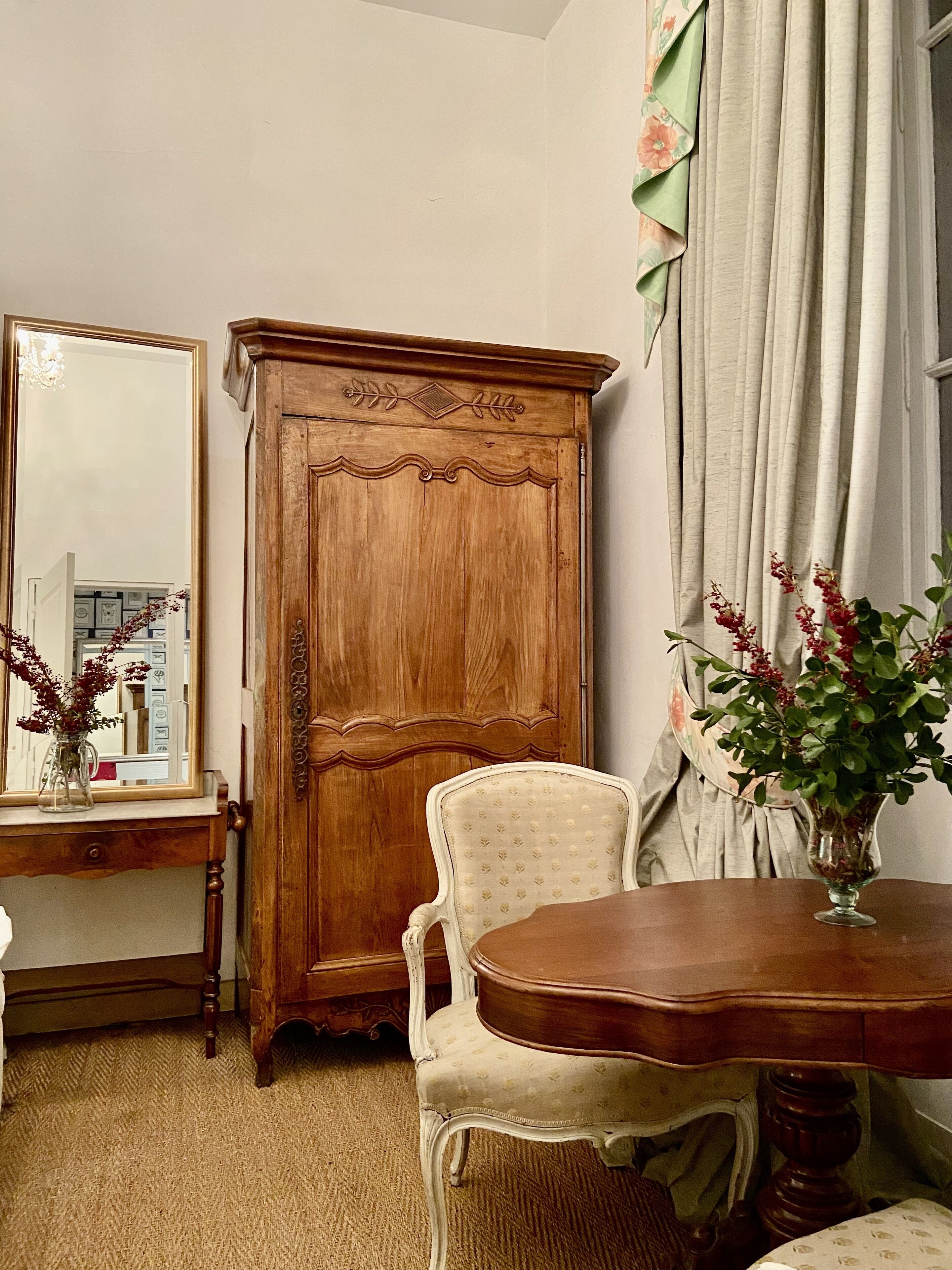 A room with a wooden wardrobe, a mirror, a floral chair, and a table with a vase of flowers.
