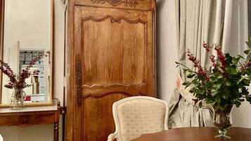 A room with a wooden wardrobe, a mirror, a floral chair, and a table with a vase of flowers.