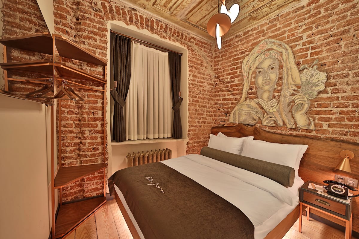 Een slaapkamer met een bakstenen muur, een bed met wit en bruin beddengoed, een houten hoofdeinde, een nachtkastje met een lamp, en een plank met hangende kleding.