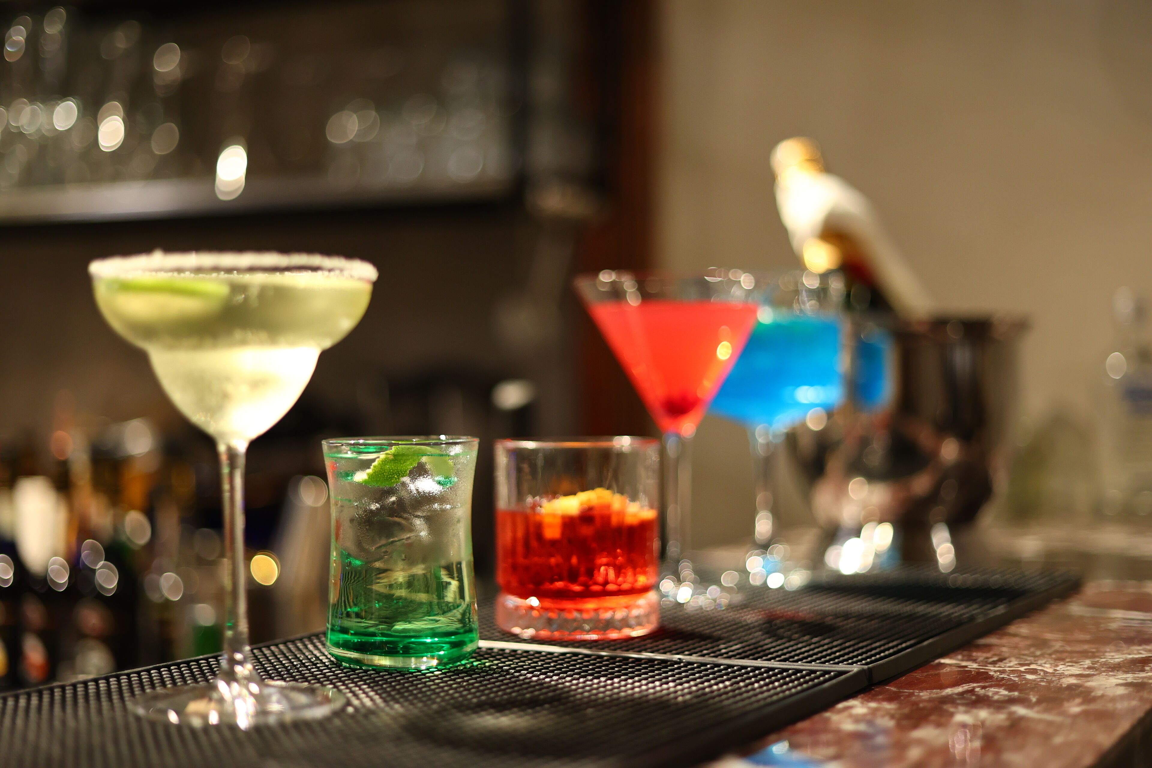A bar area with various cocktails in different glasses, a bartender's hat, and a blurred background with bottles.
