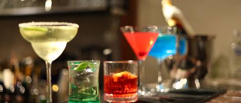 A bar area with various cocktails in different glasses, a bartender's hat, and a blurred background with bottles.