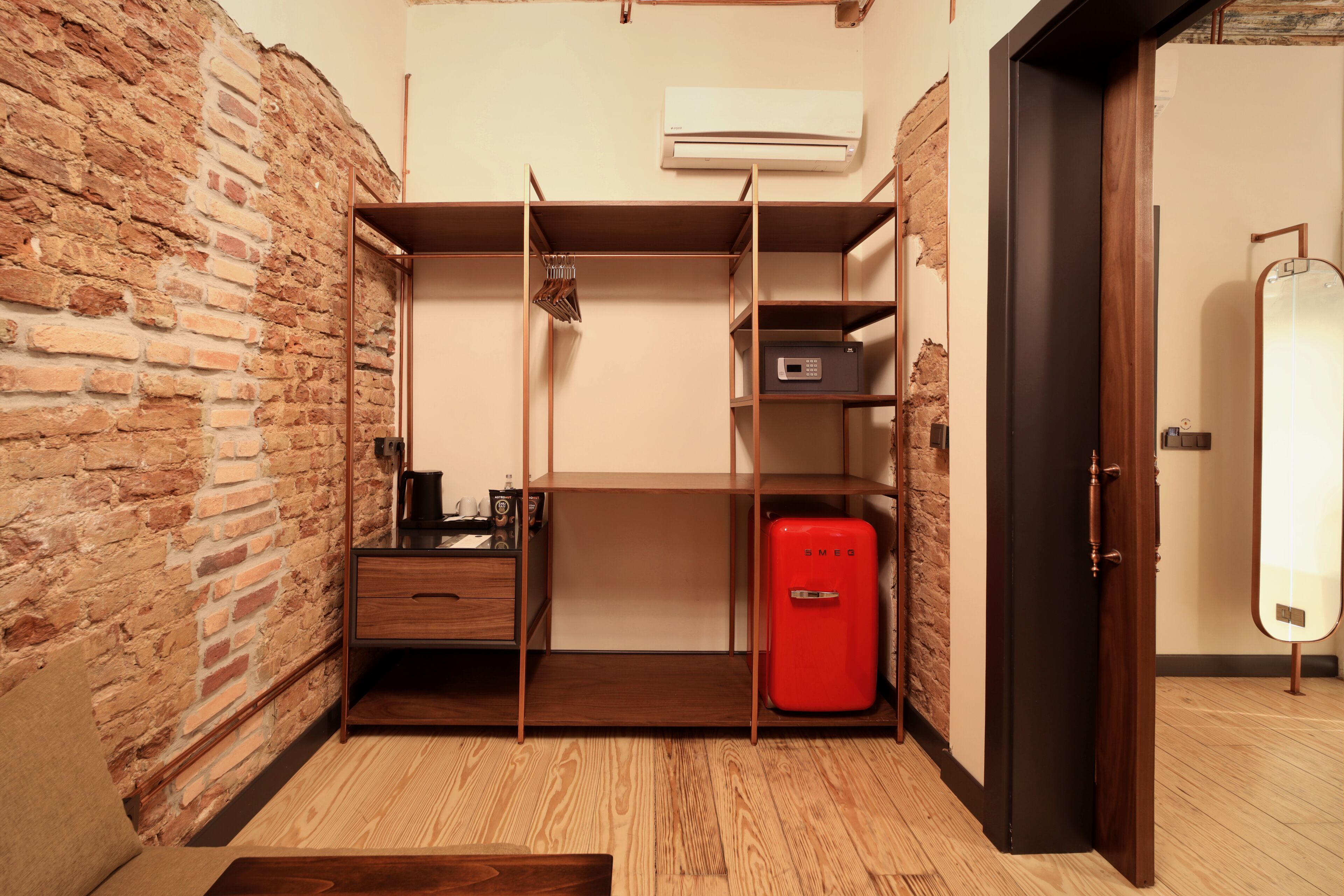 A compact room with a brick wall, a wooden floor, a wardrobe with shelves, a red appliance, and a door.
