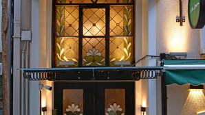 Entrada de un hotel con toldo verde, puerta de cristal con motivos florales y felpudo.