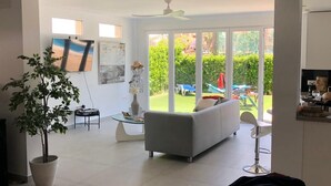 A spacious living room with a grey sofa, a dining area with a table and chairs, a large window with a view of a garden, and a ceiling fan. - Modern 3 Bed Villa - Golf Side, Own Pool, sleeps 6, Mar Menor Golf Resort     (Torre-Pacheco)
