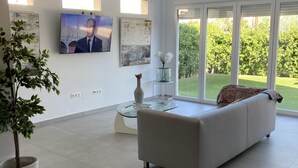 A modern living room with a sofa, a coffee table, and a TV mounted on the wall. There is a large window with a view of greenery outside. - Modern 3 Bed Villa - Golf Side, Own Pool, sleeps 6, Mar Menor Golf Resort     (Torre-Pacheco)