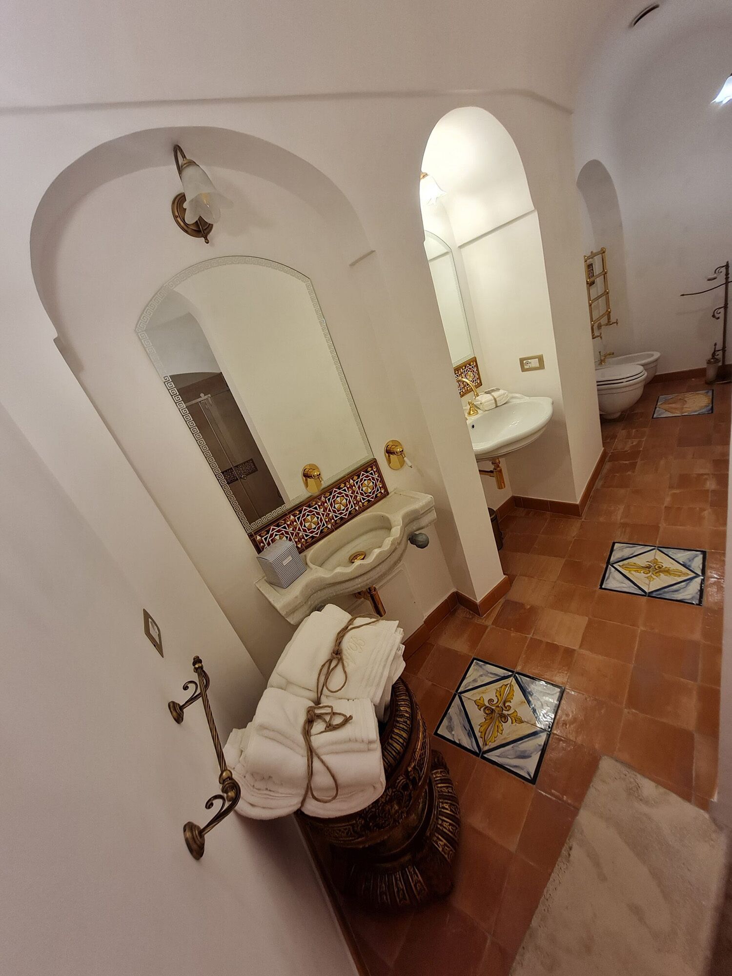 Un baño con lavamanos, espejo e inodoro. El suelo tiene azulejos decorativos y hay un perchero con toallas.