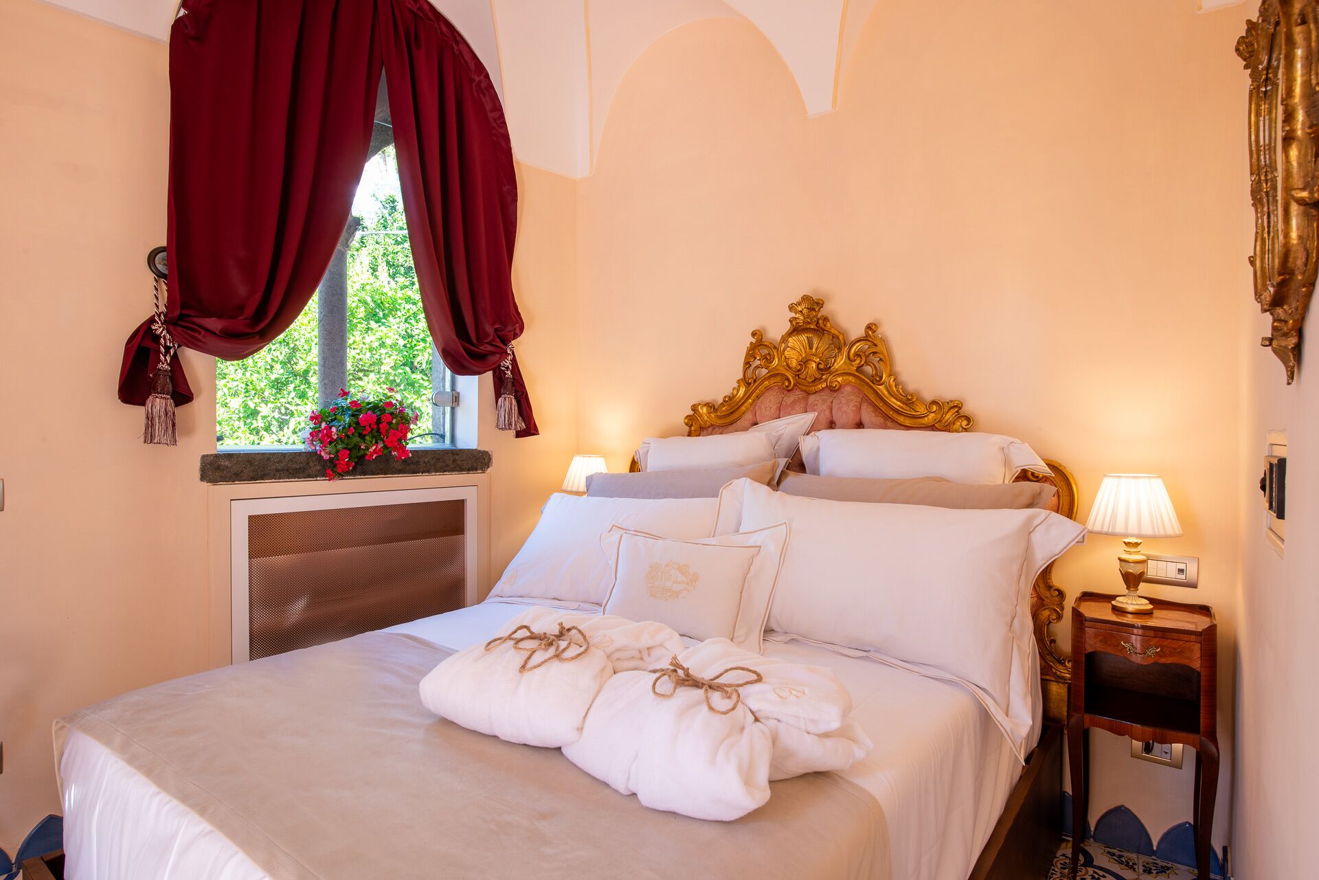 A hotel room with a large bed, a window with red curtains, a decorative headboard, and bedside lamps.
