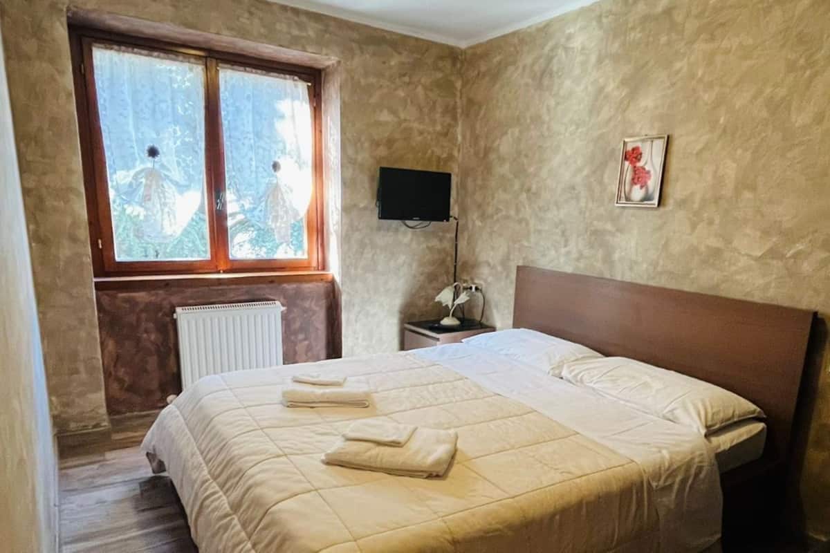 A bedroom with a large bed, a window with a view, a bedside table with a lamp, and a wall-mounted TV.