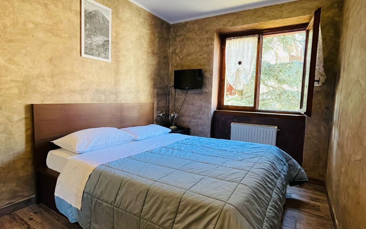 A bedroom with a bed, a window with curtains, a wall-mounted TV, and a radiator.