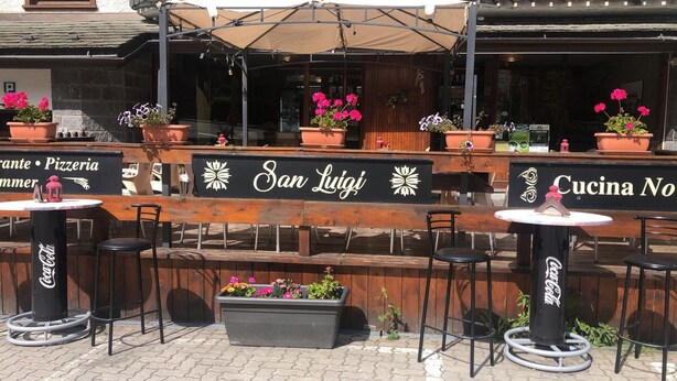 Un espace extérieur aménagé avec des tables et des chaises en bois, des plantes en pot et des panneaux indiquant « San Luigi » et « Cucina No ».