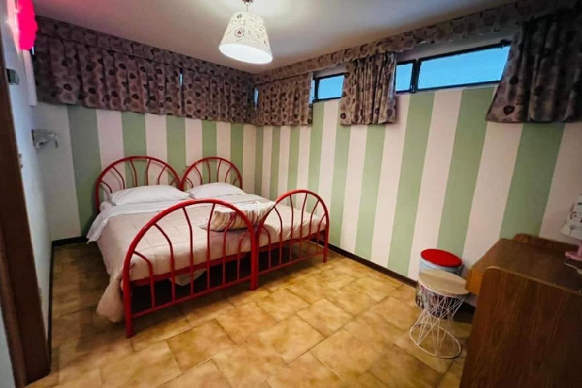 A room with a striped wall, a bed with a red metal frame, a small stool, and a wooden cabinet.