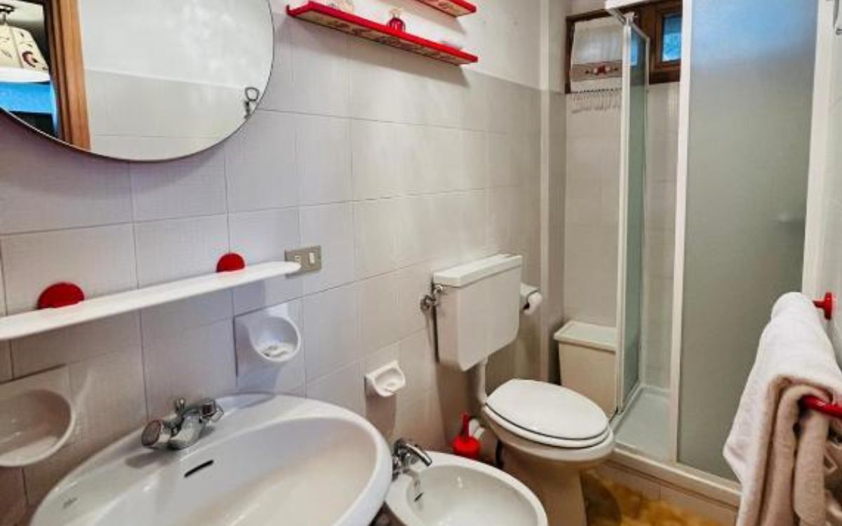 Une salle de bain équipée d’un lavabo, de toilettes et d’une douche. On y trouve des porte-serviettes rouges et un miroir au-dessus du lavabo.