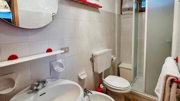 Une salle de bain équipée d’un lavabo, de toilettes et d’une douche. On y trouve des porte-serviettes rouges et un miroir au-dessus du lavabo.