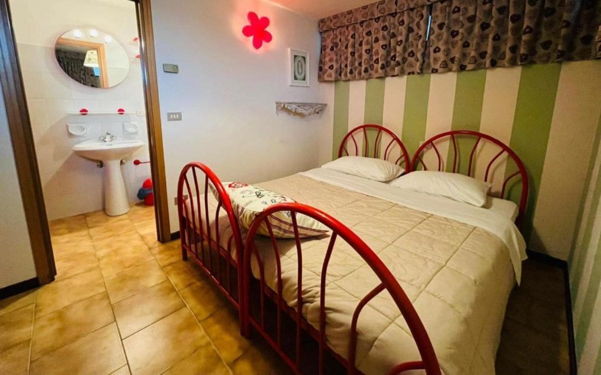 A bedroom with a single bed, a red metal frame, a white bedspread, and a red flower decoration on the wall.