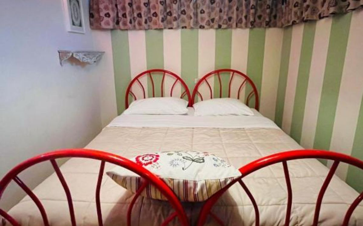 Un lit simple avec un cadre en métal rouge, une literie blanche et un coussin à motifs floraux. La pièce a des murs rayés verts et blancs et une fenêtre avec un rideau à motifs.