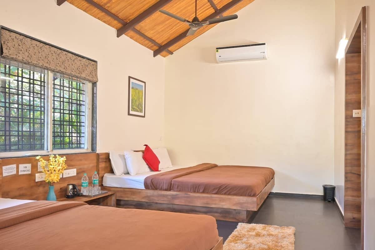 A hotel room with two beds, a ceiling fan, an air conditioning unit, a window with a view, and a bedside table with a vase of flowers.