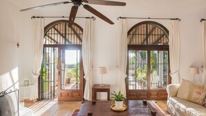 A room with a ceiling fan, two large arched windows with curtains, a wooden coffee table with a plant, a sofa, and a small table with a lamp. - Exceptional private villa with sea view & heated pool (Mijas)